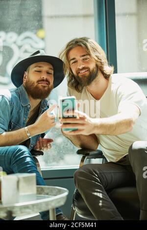 Handsome man in black hat sitting and enjoying process of taking selfie on mobile phone with friend in cafe Stock Photo