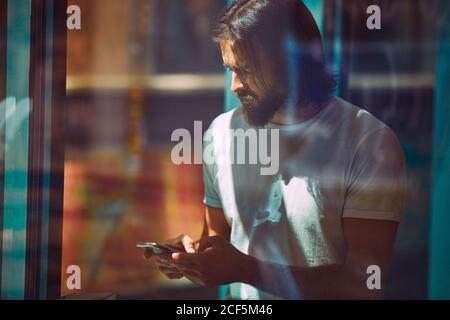 Young bearded handsome man leaning on wall with mobile phone in hands thoughtfully texting Stock Photo