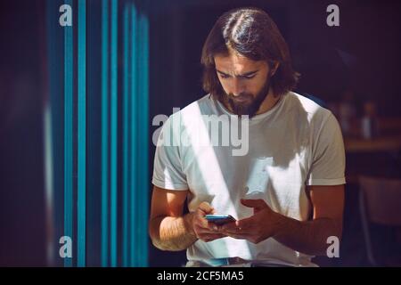 Young bearded handsome man leaning on wall with mobile phone in hands thoughtfully texting Stock Photo