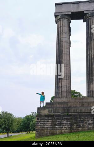Side view of Woman in colorful jacket standing on memorial monument steps and pointing with finger at sky on sunny day in park Stock Photo