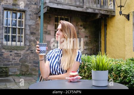 Dreamy female in casual wear taking hot drink from disposable cup while sitting at round table with cupcake garnished with buttercream and raspberries in street cafeteria in sunlight Stock Photo