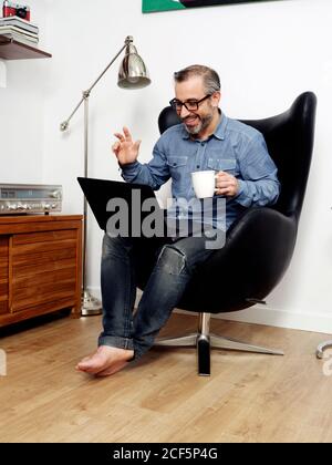 man working on laptop and talking on video conference smiling having relaxed coffee on couch at home Stock Photo