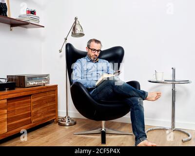 man reading a book having a relaxed coffee in the armchair at home Stock Photo