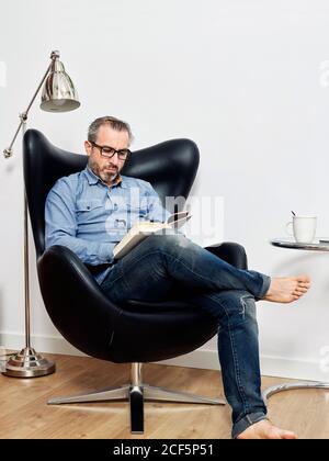 man reading a book having a relaxed coffee in the armchair at home Stock Photo