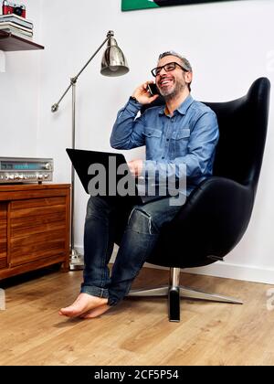 man working on the laptop and talking on the mobile phone smiling having a relaxed coffee in the armchair at home Stock Photo