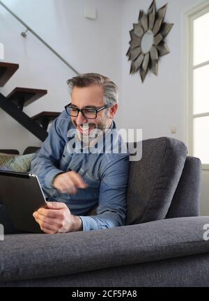 Smiling man using tablet surfing the internet alone on the couch in the living room at home Stock Photo