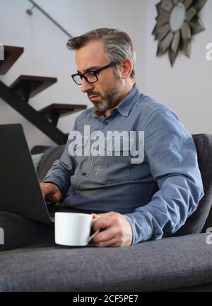 Serious man using laptop surfing the internet alone on the couch in the living room at home with coffee Stock Photo