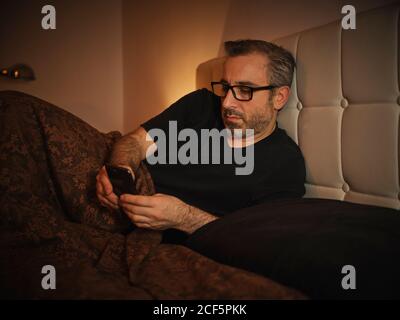 Relaxed and serious man in bed reading on the internet with a smartphone before sleeping and resting Stock Photo