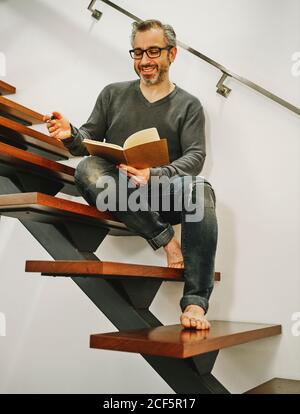 Low angle of happy mature male in eyeglasses relaxing with cup of coffee on wooden steps in contemporary apartment and writing plans in notebook Stock Photo