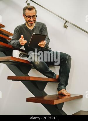 Happy man in casual clothing sitting on wooden stairs using tablet during rest in modern home Stock Photo