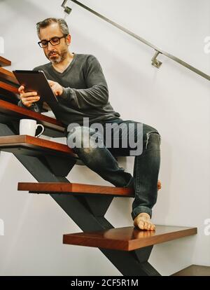 Happy man in casual clothing sitting on wooden stairs using tablet during rest in modern home Stock Photo