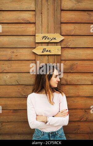 Confident beautiful Woman with hands crossed leaning on wooden wall with two signs looking away in Tarifa, Spain Stock Photo