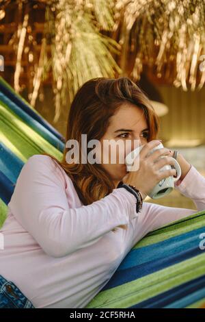 Crop Calm female in casual outfit sitting in hammock drinking mug of hot drink in courtyard and enjoying summer holiday Stock Photo