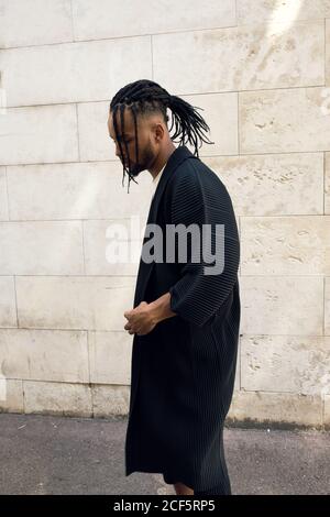 Fashionable young African American male in stylish outfit posing in street near grungy wall on sunny day on city street Stock Photo