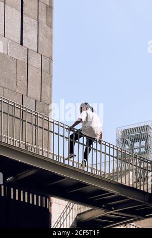 Young African American man sitting on railing of bridge while spending time on city street on sunny day Stock Photo