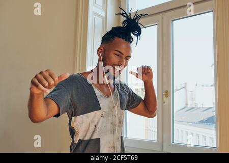 Smiling African American man with braids dancing to music in earphones at home on window background Stock Photo