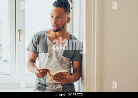 Young African American man with mohawk pigtails standing leaning on windowsill and reading book indoors Stock Photo