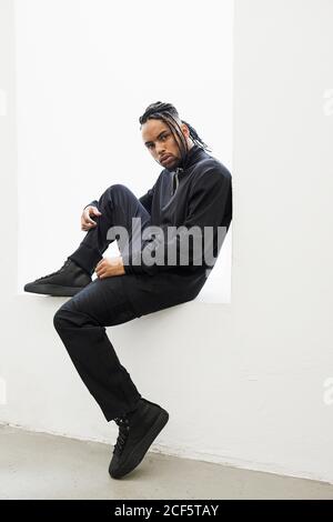 Side view of African American man in black clothes with braided hair sitting isolated on white background Stock Photo