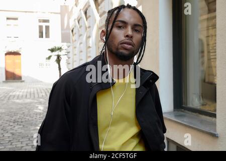 Serious young man with braids and earphones and mobile phone standing and looking at camera on urban background Stock Photo