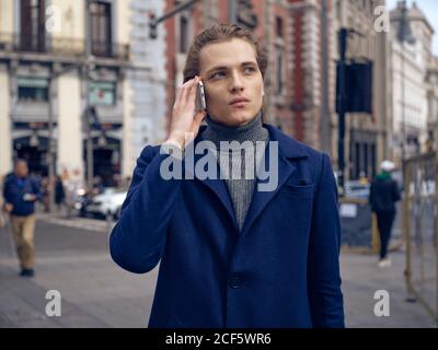 Modern elegant male in trendy coat looking away pensively while having phone conversation on city street Stock Photo