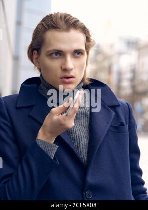 of serious young businessman in elegant coat using mobile phone while standing on stone staircase on city street Stock Photo