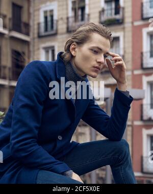 Low angle side view of serious elegant young male in stylish coat leaning on hand and thinking while sitting on street against blurred building Stock Photo