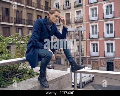 Low angle side view of serious elegant young male in stylish coat leaning on hand and thinking while sitting on street against blurred building Stock Photo