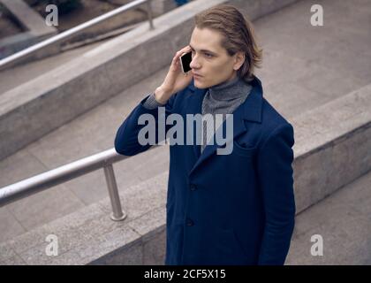 From above of serious young businessman in elegant coat checking message on mobile phone while standing on stone staircase on city street Stock Photo