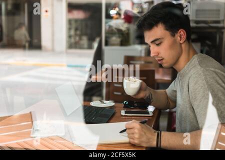 Side view of young man with cup of coffee writing in notepad while sitting at table behind glass and studying in cafeteria Stock Photo