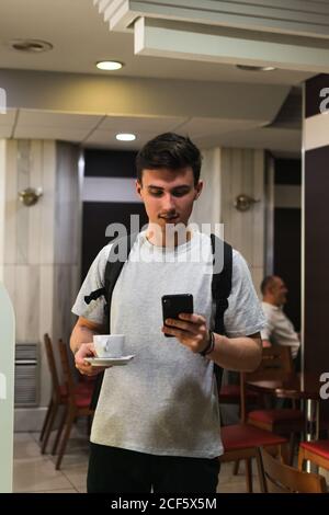 Young man with backpack carrying cup of hot beverage and browsing smartphone while standing in cafeteria Stock Photo