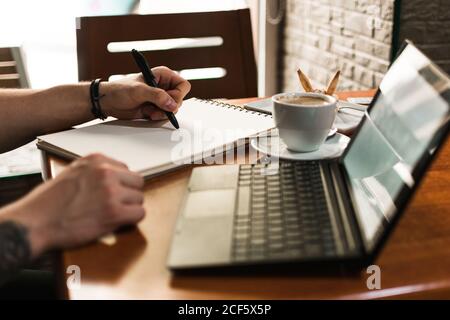 unrecognizable man sketching in notepad near cup of coffee and laptop while sitting at table an working on remote project in cafeteria Stock Photo