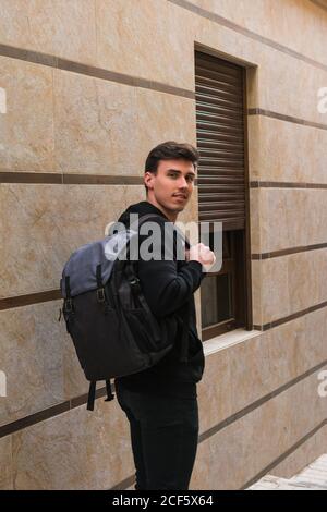 Side view of positive young man with backpack looking at camera while standing near window of modern building on city street Stock Photo