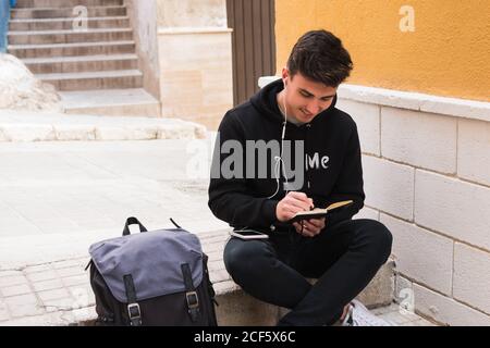 Cheerful young man in casual clothes listening to music in earphones and writing in notebook while sitting on step near backpack on city street Stock Photo