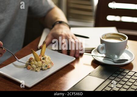 unrecognizable man sitting at table with laptop and cup of coffee and eating tasty salad in restaurant Stock Photo