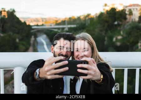 Joyful young multiracial couple in casual wear taking selfie on mobile phone while standing together on bridge with green trees and city buildings in background Stock Photo