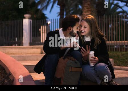 Cheerful multiethnic couple in trendy warm clothes drinking takeaway coffee and browsing smartphone while sitting together in tropical park in evening Stock Photo