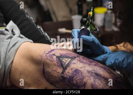 Crop unrecognizable tattoo master in gloves using professional machine while creating picture on male leg in salon Stock Photo