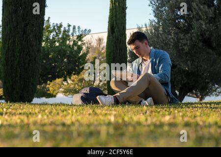 Smart concentrated man with backpack studying writing on notepad while sitting in park grass with crossed legs in sunny day Stock Photo