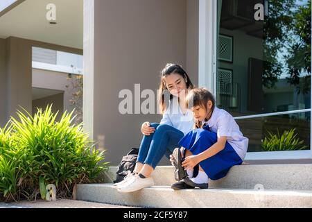 Asian mother looking her daughter primary students in uniform to wearing their own shoes infront of home in the morning school routine for day in the Stock Photo