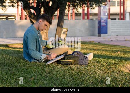 Smart concentrated man with backpack studying at laptop and writing on a notepad sitting in park grass with crossed legs in sunny day Stock Photo