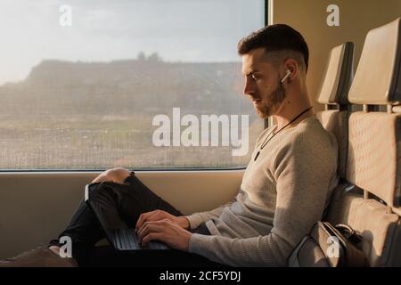 Side view of young man in wireless earphones typing on laptop during journey in subway in sunny day Stock Photo