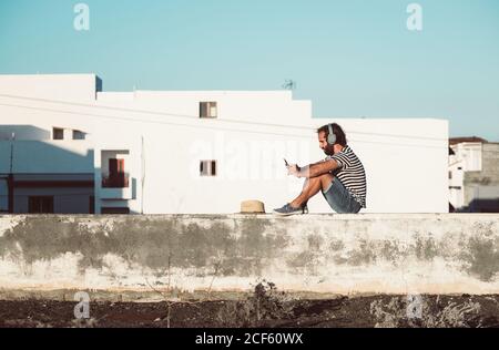 Side view of man listening to music and using mobile phone while relaxing on embankment in La Restinga Stock Photo