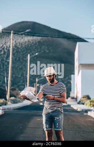 Young trendy man searching destination on map while walking on road against mountain background in La Restinga, El Hierro Stock Photo
