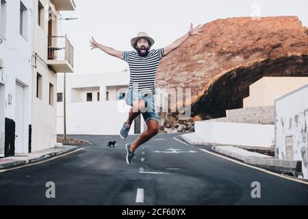 Stylish bearded man jumping for joy with outstretched arms and black cat on road in La Restinga, El Hierro Stock Photo