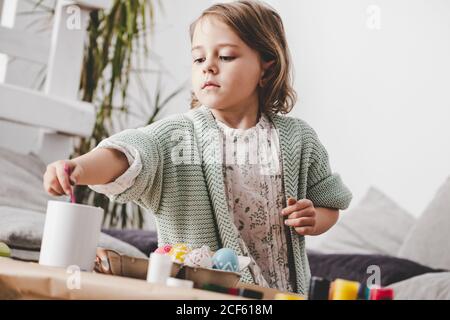 girl with brush coloring chicken eggs at table in room Stock Photo