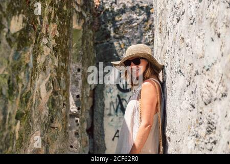 Tanned stylish Woman in hat, sunglasses and light costume enjoying sightseeing leaning in ancient stone wall looking at camera Stock Photo