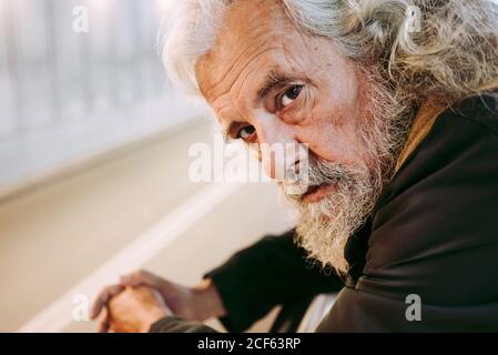 Bearded male tourist with grey hair in outerwear leaning on metal railing construction while standing under bridge with backpack looking at camera Stock Photo