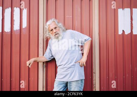 Mature aged man with grey hair in gray cotton shirt and jeans leaning on red metal wall while resting hand with signet on waist Stock Photo