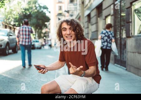 Cheerful casual surprised man making faces while on smartphone while sitting on baluster on city street Stock Photo