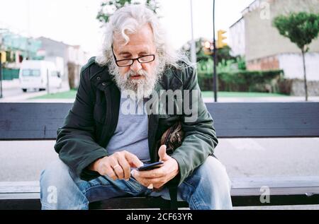 Concentrated grey bearded man in casual clothes wearing eyeglasses and messaging on mobile phone while resting on street bench Stock Photo